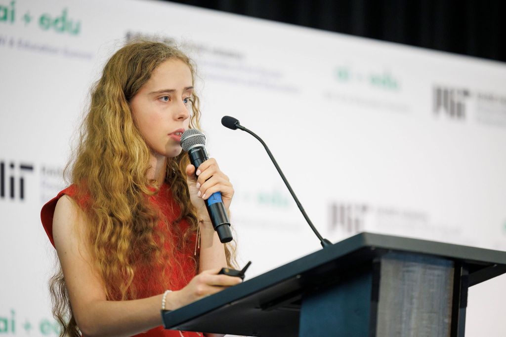 Teenage girl with curly brown hair and red blouse speaks at a podium